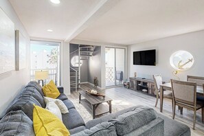 A modern living room with a grey sofa, yellow cushions, a wooden coffee table, a TV mounted on the wall, and a staircase leading to a balcony. - Solare Garden Villa 679F (South Padre Island)