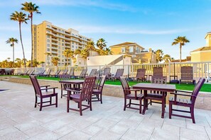 An outdoor seating area with tables and chairs, surrounded by a white fence, with palm trees and buildings in the background. - Solare Garden Villa 679F (South Padre Island)