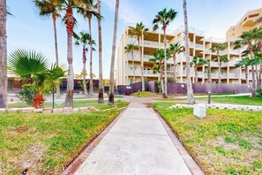 A pathway leading to a multi-story building with balconies, surrounded by palm trees and a well-maintained lawn. - Solare Garden Villa 679F (South Padre Island)