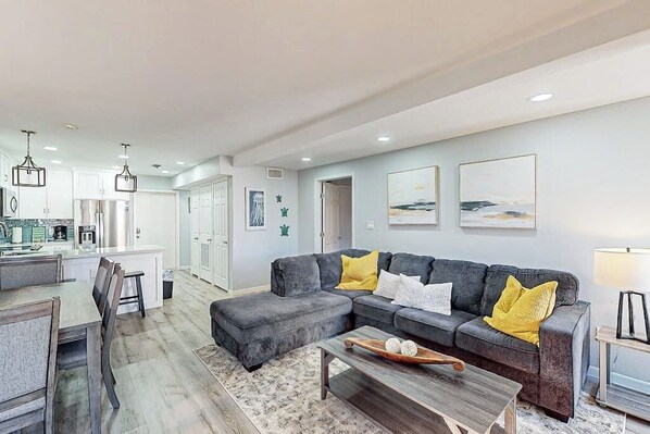 A modern living room with a grey sectional sofa, a wooden coffee table, and a kitchen in the background. - Solare Garden Villa 679F (South Padre Island)