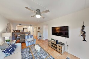 A modern living room with a flat-screen TV, a ceiling fan, and a dining area with a kitchen in the background. - Surfside I Condominiums #301 (South Padre Island)