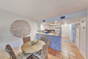 A dining area with a round table and wicker chairs, a kitchen in the background, and a tiled floor. - Surfside I Condominiums #301 (South Padre Island)