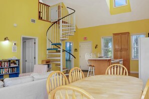 Ein Zimmer mit Wendeltreppe, einem Essbereich mit Holzsesseln, einer Küche mit Kühlschrank und einem Bücherregal.