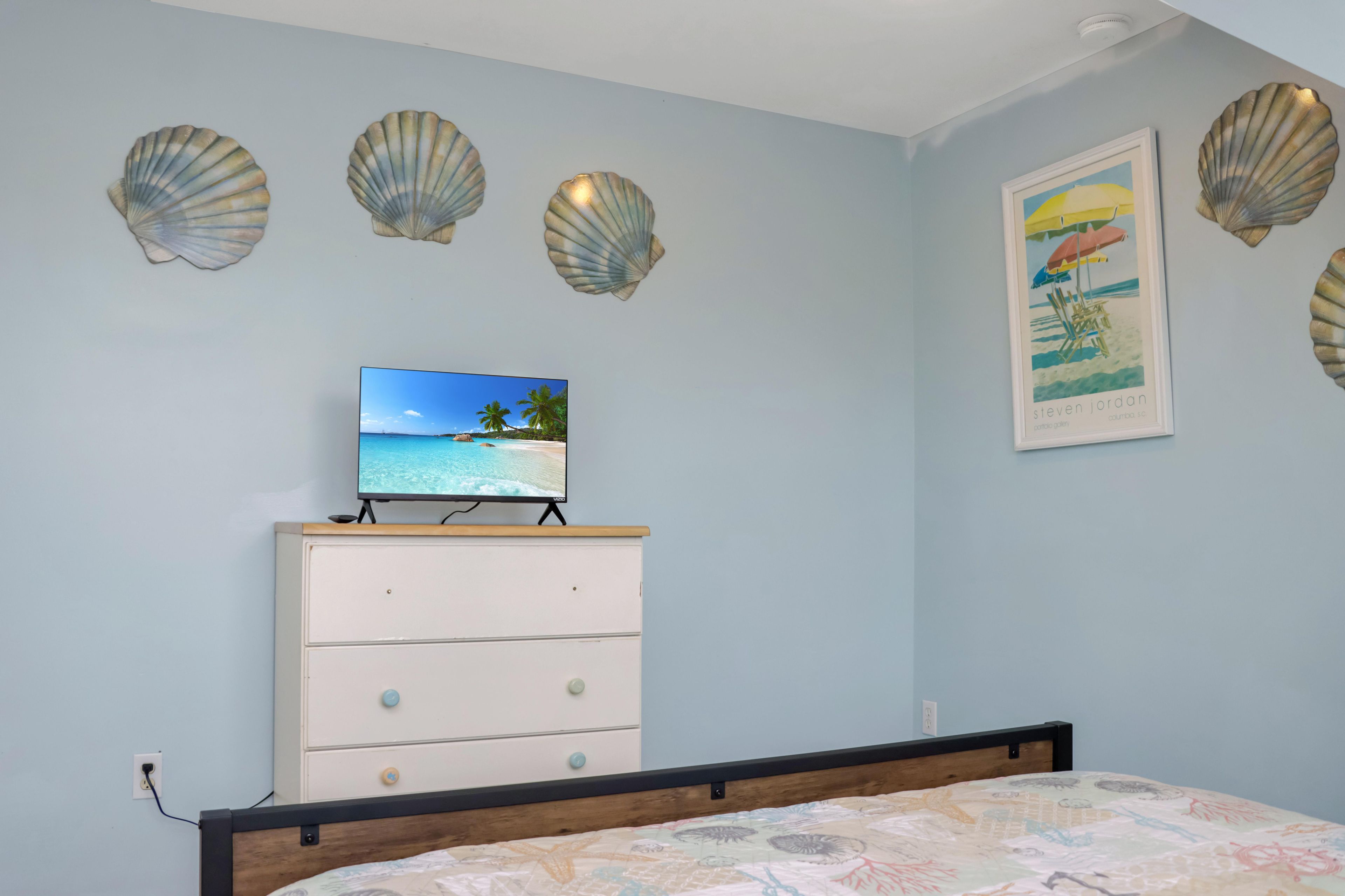 Quarto com cama, penteadeira, televisão exibindo uma cena de praia e conchas decorativas na parede.
