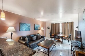 A modern living room with a sofa, coffee table, dining table, and chairs. There are two wall-mounted lamps and a television on a wooden stand. - Gulfview I #312 - Ocean View & Hot Tub (South Padre Island)