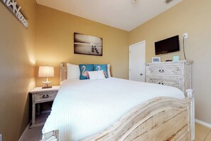 A bedroom with a bed, nightstand, lamp, artwork, television, and a dresser. - Kingfisher - Shared Pool, Walk to the Beach (South Padre Island)