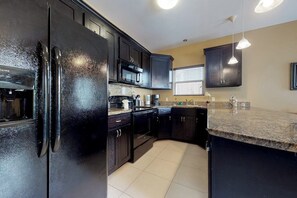 A kitchen with dark cabinets, a black refrigerator, a microwave, and a granite countertop. - Kingfisher - Shared Pool, Walk to the Beach (South Padre Island)