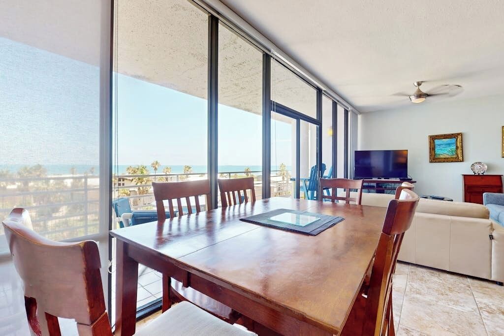 A spacious room with a large dining table, chairs, a television, and a view of the ocean.
