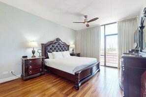 Ein Schlafzimmer mit einem großen Bett, Holzboden, Deckenventilator und Blick ins Freie durch ein Fenster.