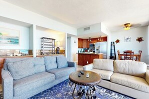 Un salon avec un canapé gris, un canapé beige et une table basse. Au fond, on aperçoit une cuisine avec des meubles en bois et un four à micro-ondes.