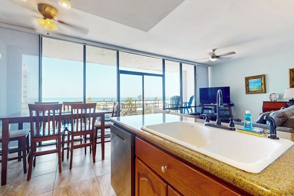 A kitchen with a large window, a dining area with chairs, a TV, and a ceiling fan.