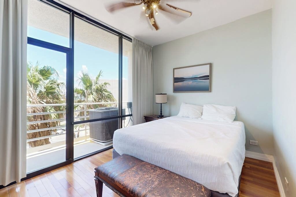Ein Hotelzimmer mit einem großen Bett, einem Deckenventilator, einem Balkon mit Blick auf Palmen und einem Bild an der Wand.