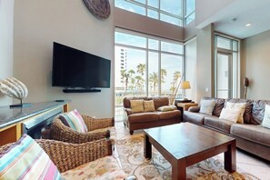 A spacious living room with a flat-screen TV, comfortable seating, and large windows offering a view of palm trees. - Sapphire Condominiums Bungalow #106 (South Padre Island)