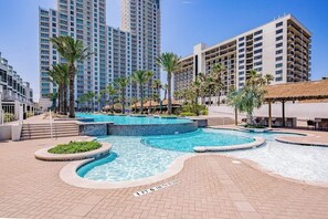 A hotel pool area with multiple swimming pools, palm trees, and a thatched-roof structure. - Sapphire Condominiums Bungalow #106 (South Padre Island)