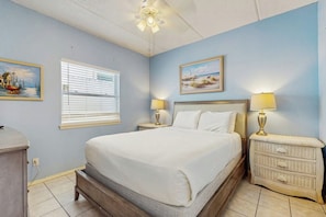A bedroom with a bed, two bedside tables with lamps, a window with blinds, and two wall-mounted paintings. - Coronado Condominiums #102 (South Padre Island)