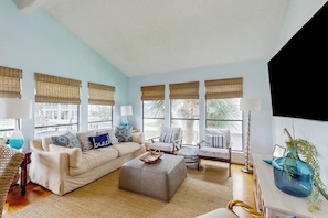 A spacious living room with a beige sofa, a grey ottoman, and striped armchairs. Large windows with bamboo blinds allow natural light. - Piper's Place - Short Walk to Beach, Dogs OK (Galveston)