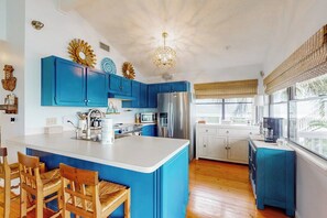 A kitchen with blue cabinets, a central island, wooden chairs, and a large window with blinds. - Piper's Place - Short Walk to Beach, Dogs OK (Galveston)