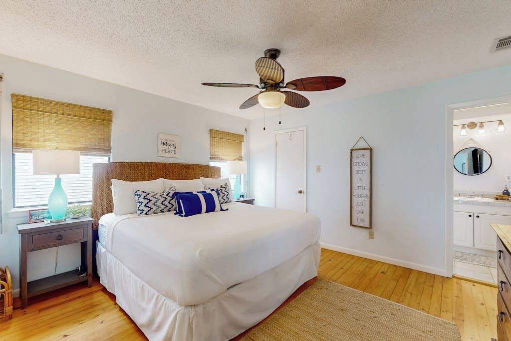 A bedroom with a bed, a ceiling fan, a bedside table, a lamp, a window with blinds, a wall-mounted mirror, and a wall with a motivational quote.