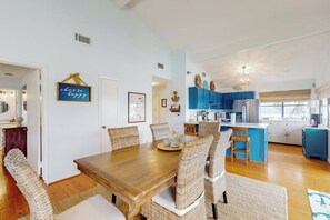 A dining area with a wooden table and wicker chairs, a kitchen with blue cabinets, and a sign on the wall. - Piper's Place - Short Walk to Beach, Dogs OK (Galveston)