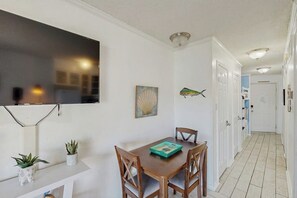 A dining area with a table, chairs, a flat-screen TV mounted on the wall, a potted plant, and a framed artwork. - Seawall Treasure - Heated Pool & Private Balcony (Galveston)