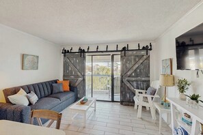 Een woonkamer met een bank, salontafel en stoelen, een balkon met een schuifdeur, en een televisie aan de muur.