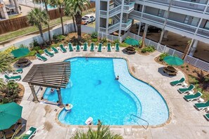 A swimming pool with lounge chairs and umbrellas, surrounded by a fence and palm trees. - Seawall Treasure - Heated Pool & Private Balcony (Galveston)