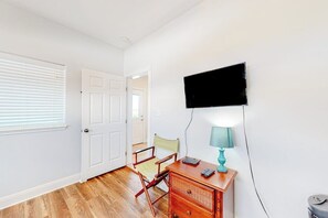 A room with a wooden floor, a desk with drawers, a chair, a television mounted on the wall, and a window with blinds. - Another Day In Paradise (Port Aransas)