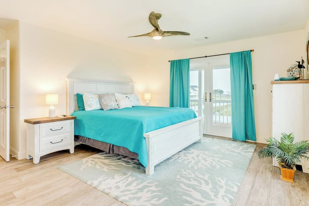 A bedroom with a bed, nightstands, a ceiling fan, a window with curtains, and a view outside.