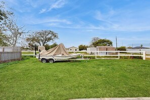 A trailer with a boat covered by a tarp in a grassy area with a white fence and buildings in the background. - The Saltbox - Private Pool, Fire Pit & Yard (Corpus Christi)