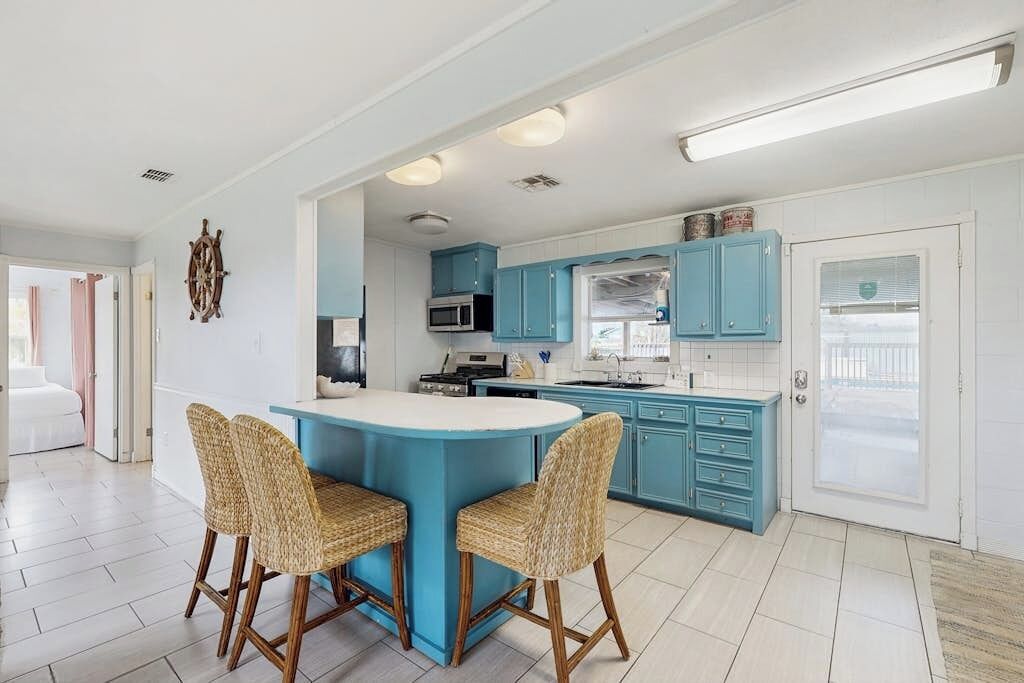 Una cocina con una isla central, dos sillas de mimbre, armarios color turquesa y una encimera blanca.