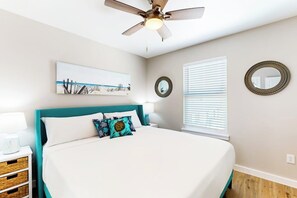 A bedroom with a bed, a ceiling fan, a window with blinds, a wall-mounted mirror, and a painting. - Classy Bum - Covered Patio & Alfresco Dining (Port Aransas)
