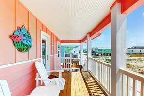 A covered porch with white railings, a wooden deck, and a view of a beach and buildings in the background. - Classy Bum - Covered Patio & Alfresco Dining (Port Aransas)