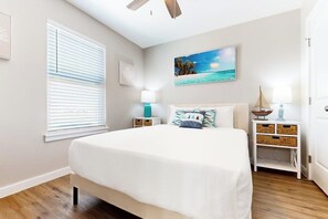 A bedroom with a bed, two bedside tables, a ceiling fan, a window with blinds, and a painting on the wall. - Classy Bum - Covered Patio & Alfresco Dining (Port Aransas)