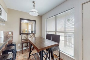 A dining area with a wooden table and chairs, a hanging light fixture, and a window with blinds. - Pirate's Crossing #B6 (Corpus Christi)