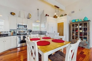 A modern kitchen with white cabinetry, a wooden dining table with red placemats, and a rustic cabinet with glass doors. - The Purple Beach House (Rockport)