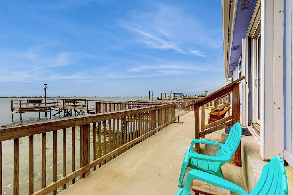 A wooden pier extending into a body of water, with a building on the right side and turquoise chairs in the foreground.