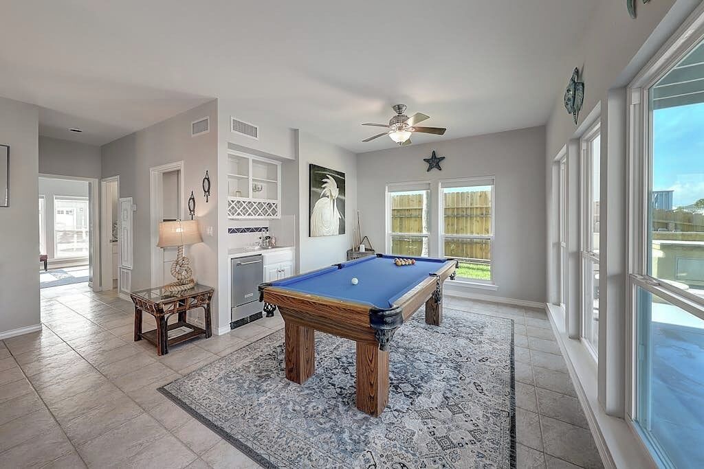 A pool table in a room with a ceiling fan, a lamp, and a view through large windows.