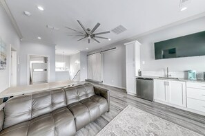 A modern living room with a leather sofa, a ceiling fan, recessed lighting, a kitchen area with white cabinets, and a flat-screen TV mounted on the wall. - Inkies Retreat - Waterfront Home with Swim Spa (Rockport)