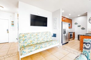 A modern living room with a sofa, a flat-screen TV, and a kitchen area with wooden cabinets and a refrigerator. - Pointe West Resort - Peaceful at the Pointe (Galveston)