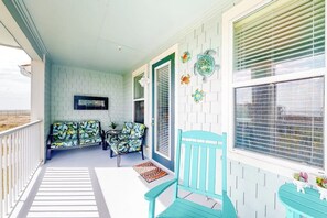 A covered porch with a white railing, a blue bench, a table with a vase of flowers, and a wall decorated with colorful plates. - Pointe West Resort - Peaceful at the Pointe (Galveston)
