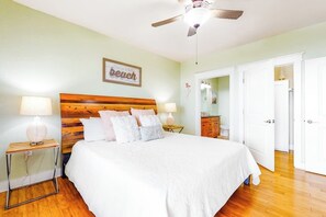 A bedroom with a wooden headboard, a bed with white bedding, two bedside lamps, a ceiling fan, and a door leading to another room. - Pointe West Resort - Peaceful at the Pointe (Galveston)