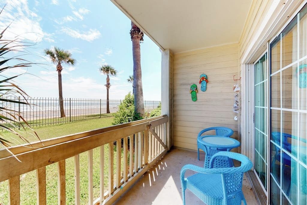 A balcony with a wooden railing, two blue chairs, and a view of the beach with palm trees.