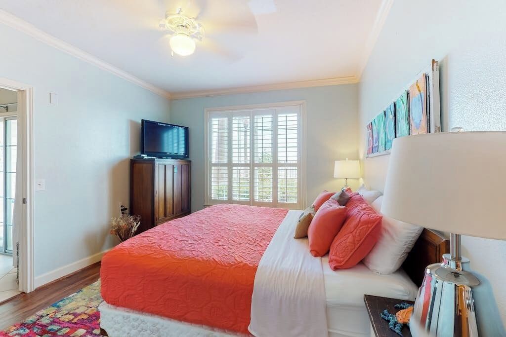 A bedroom with a large bed, a television, a window with blinds, and a lamp.