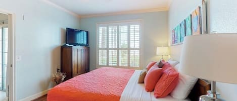 A bedroom with a large bed, a television, a window with blinds, and a lamp.