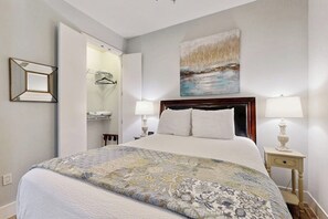 A hotel room with a bed, two bedside lamps, a painting on the wall, and a mirror with a hanging garment. - Infinity Sea of Serenity (Galveston)
