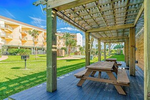 An outdoor wooden deck with picnic tables, surrounded by a well-maintained lawn and a building with balconies in the background. - Seahab, Maravilla Resort, Pools, Hot Tub, Beach (Galveston)