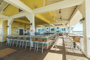 An outdoor bar area with multiple seating options, including bar stools and tables, under a covered structure with a view of the sea. - Pointe West Resort - OceanScape (Galveston)