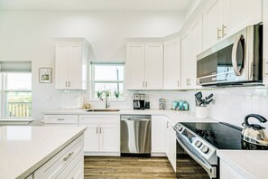 A modern kitchen with white cabinetry, stainless steel appliances, and a window providing natural light. - Vacationers Valhalla (Galveston)