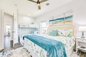 Une chambre avec des lits superposés, un ventilateur de plafond, un lit recouvert d’une couverture bleue, une table de chevet avec une lampe et des œuvres d’art accrochées au mur.