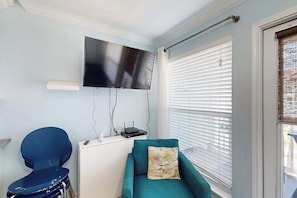 A room with a flat-screen TV mounted on the wall, a teal sofa with a floral cushion, a blue chair, and a white desk with a router. - Seascape Condos #1219 (Galveston)